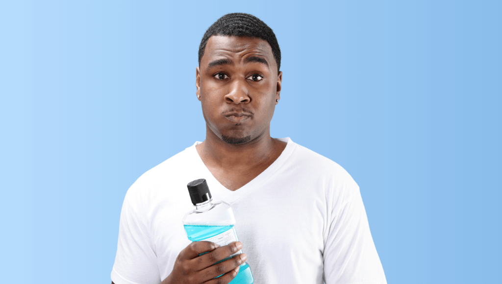 Jeune homme portant un t-shirt blanc tenant une bouteille de bain de bouche bleu et ayant une expression de doute sur fond bleu Afbeelding