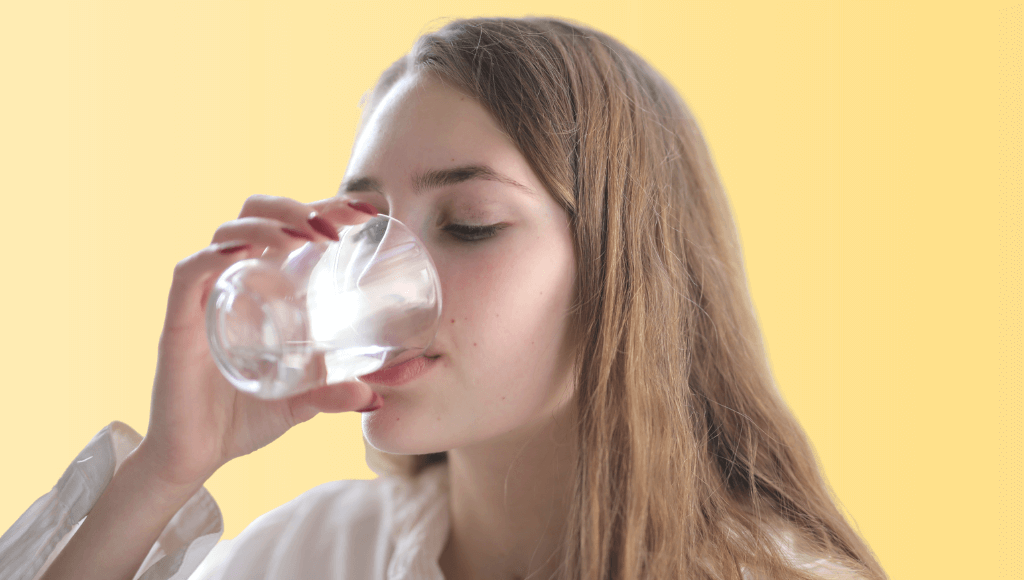 Jeune femme brune aux cheveux longs de profil buvant un verre d'eau sur fond jaune Immagine