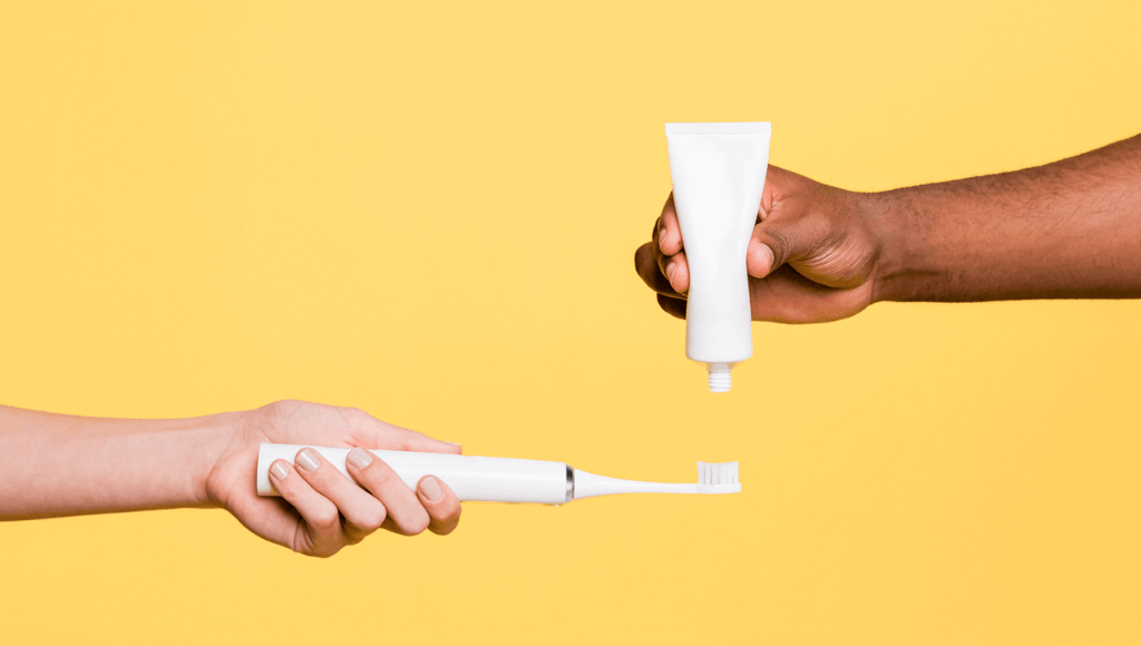 Main d'homme noir mettant du dentifrice blanc sur une brosse à dents électrique sonique blanche tenue par une main de femme blanche sur fond jaune Afbeelding