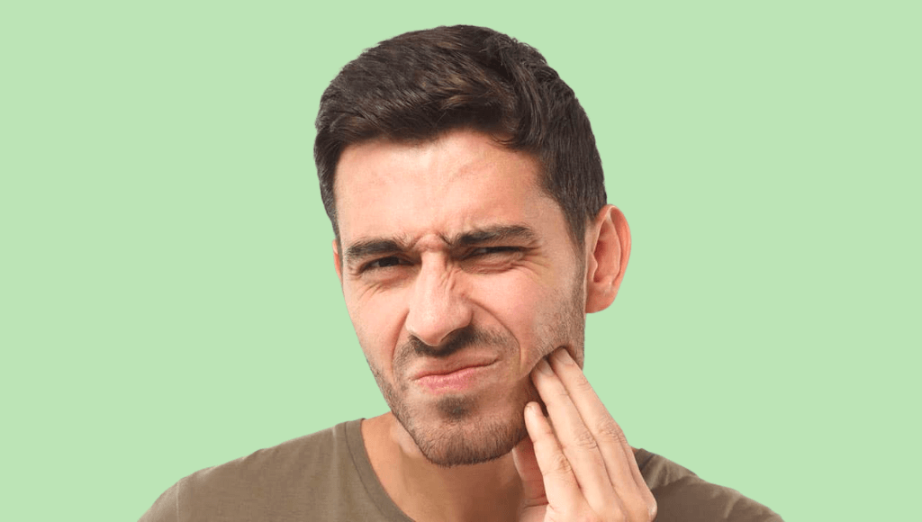 Homme aux cheveux bruns faisant une grimace en se touchant la mâchoire avec la main gauche sur fond vert Immagine